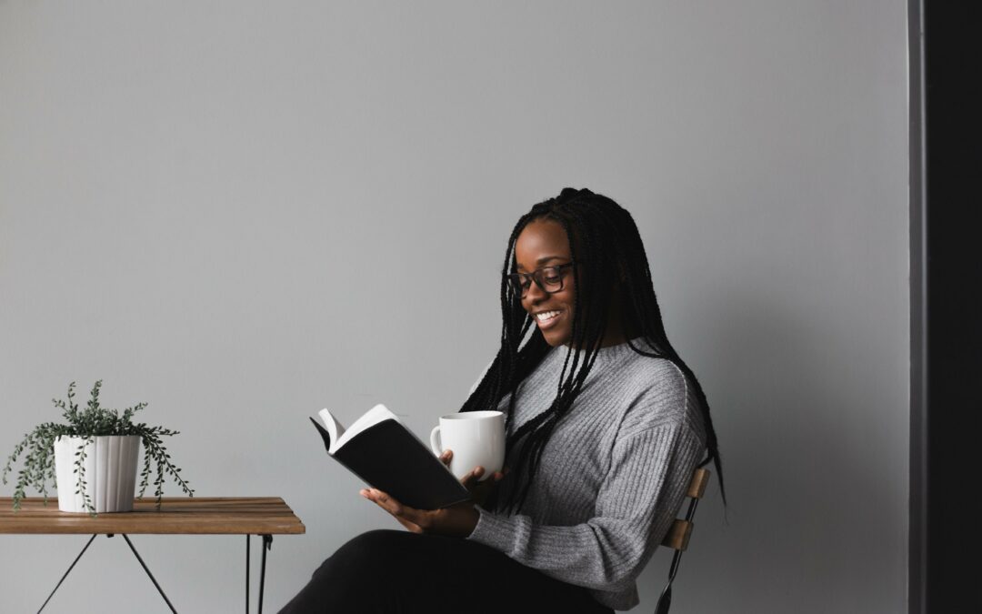 african american woman sitting at table, reading a book and holding a cup of coffee